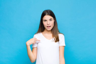 Genç ve güzel bir kadın ağzı açık, şaşkın ve şaşırmış görünüyor, mavi duvara karşı kendini işaret ederek.
