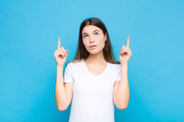 Genç ve güzel bir kadın huşu içinde ve ağzı açık bir şekilde mavi duvara karşı şaşkın ve şaşkın bir bakışla yukarıya bakıyor.