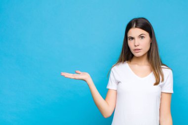 Genç ve güzel bir kadın şaşırmış ve şoka uğramış görünüyor, çenesi düşmüş, mavi duvara karşı eli açık bir obje tutuyor.