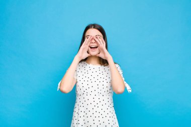 Genç, güzel bir kadın mutlu, heyecanlı ve pozitif hissediyor. Elleri ağızda, mavi duvara doğru bağırıyor.