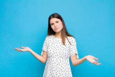 Şaşkın, şaşkın ve stresli görünen genç bir kadın, farklı seçenekler arasında merak ediyor, mavi duvarda belirsizlik hissediyor.