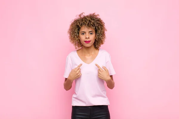 年轻的非洲裔美国女人带着困惑和好奇的神情指着自己 被选在粉红的墙上 感到震惊和惊奇 — 图库照片