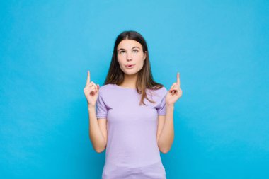 Genç ve güzel bir kadın huşu içinde ve ağzı açık bir şekilde mavi duvara karşı şaşkın ve şaşkın bir bakışla yukarıya bakıyor.