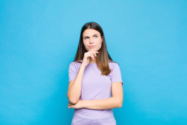 Genç, güzel bir kadın düşünüyor, kuşkulu ve şaşkın hissediyor, farklı seçenekleri var, mavi duvara karşı hangi kararı vereceğini merak ediyor.