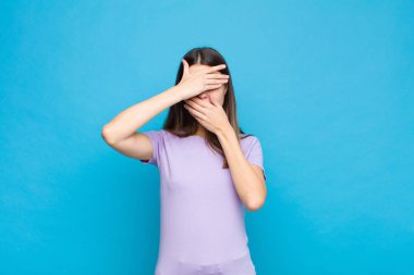 Genç ve güzel bir kadın iki eliyle kameraya hayır diyerek yüzünü kapatıyor! Resimleri reddetmek ya da mavi duvara karşı fotoğraf yasaklamak