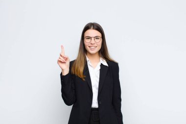 Genç iş kadını mutlu bir şekilde gülümsüyor, bir eliyle beyaz duvara uzayı gösteriyor.