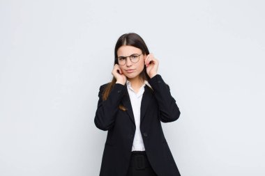 Genç iş kadını kızgın, stresli ve sinirli görünüyor, her iki kulağı da sağır edici bir gürültü, ses ya da beyaz duvara karşı yüksek sesle müzik
