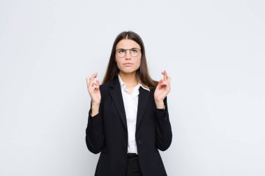 Genç iş kadını endişeyle dua ediyor ve beyaz duvara karşı endişeli bir bakışla iyi şanslar diliyor.