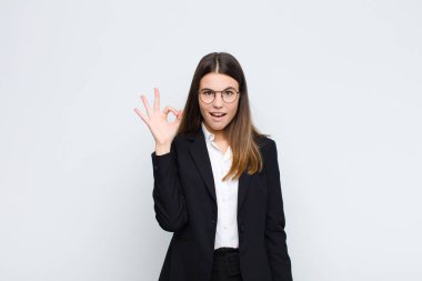 Genç iş kadını kendini başarılı ve tatmin olmuş hissediyor, ağzı açık gülümsüyor, beyaz duvara karşı elle işaret veriyor.