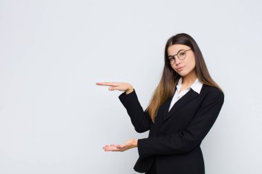 Genç iş kadını, iki eli de yan tarafında olan bir objeyi tutuyor. Beyaz duvara karşı bir objeyi gösteriyor, sunuyor ya da reklamını yapıyor.