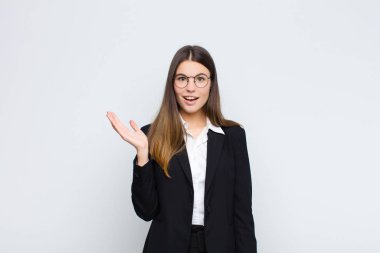 Genç iş kadını kendini mutlu, şaşırmış ve neşeli hissediyor. Pozitif bir tavırla gülümsüyor. Beyaz duvara karşı bir çözüm ya da fikir buluyor.