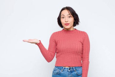 Genç ve güzel bir kadın şaşırmış ve şoka uğramış görünüyor, çenesi düşmüş, beyaz duvara karşı eli açık bir obje tutuyor.