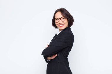 Genç iş kadını neşeyle gülümsüyor, mutlu, tatmin olmuş ve rahatlamış hissediyor, kollarını kavuşturup beyaz duvara bakıyor.