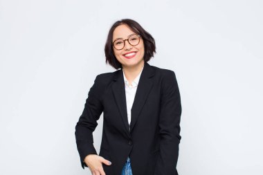 Genç iş kadını beyaz duvara karşı neşeli, mutlu, kendinden emin ve rahat bir ifadeyle gülümsüyor.