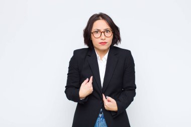 Genç iş kadını, kafası karışmış ve tuhaf bir bakışla kendini işaret ediyor. Beyaz duvara karşı seçildiği için şaşırmış ve şaşırmış.