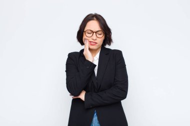 Genç iş kadını yanağını tutuyor ve acı veren diş ağrısı çekiyor, hasta, sefil ve mutsuz hissediyor, beyaz duvara karşı bir dişçi arıyor.