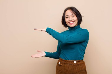 Genç ve güzel bir kadın, iki eli de kenarda olan bir objeyi tutuyor. Fotokopi alanı, bej duvara karşı bir objeyi gösteriyor, sunuyor ya da reklamını yapıyor.