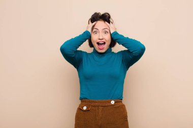 Genç ve güzel bir kadın el ele tutuşuyor, ağzı açık, son derece şanslı, şaşkın, heyecanlı ve bej rengi duvara karşı mutlu hissediyor.