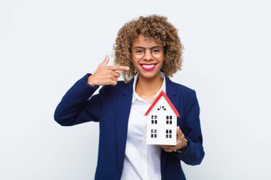 Genç Afro-Amerikan kadın, kendine güvenerek gülümsüyor. Kendi geniş gülüşünü gösteriyor. Pozitif, rahat, ev modeline karşı tatmin olmuş bir tavır.