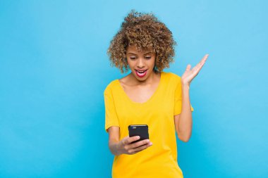 Genç Afrikalı Amerikalı mutlu, heyecanlı, şaşırmış ya da şaşırmış, gülümseyen ve hayrete düşmüş bir şekilde cep telefonuyla inanılmaz bir şey yapıyor.