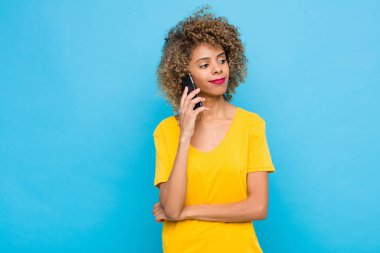 Genç Afrikalı Amerikalı mutlu bir şekilde gülümsüyor ve hayal kuruyor ya da şüphe ediyor, cep telefonuyla yan tarafa bakıyor.