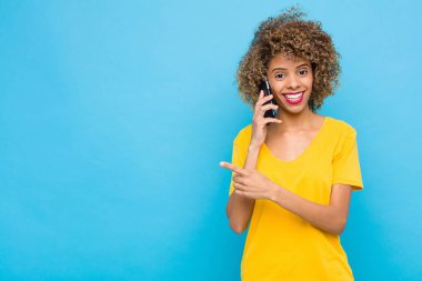 Genç Afrikalı Amerikalı neşeli bir şekilde gülümsüyor, mutlu hissediyor ve yan ve yukarı doğru işaret ediyor, fotokopi uzayında cep telefonuyla nesneleri gösteriyor.