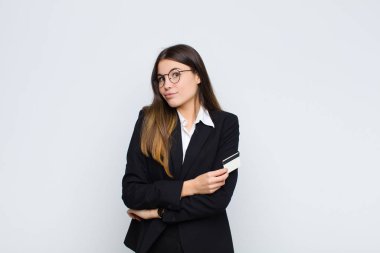 Genç, güzel bir kadın omuz silkiyor, kafası karışık ve kararsız, kolları bağlı ve kafası karışmış bir şekilde bir kredi kartıyla bakıyor.