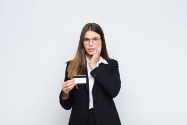 Genç ve güzel bir kadın şok olmuş ve korkmuş hissediyor, ağzı açık ve elleri kredi kartıyla yanaklarında korkmuş görünüyor.