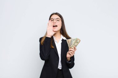 Genç, güzel bir kadın mutlu, heyecanlı ve pozitif hissediyor. Elleri ağızda bağırıyor. Banknotlarla bağırıyor.