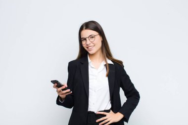 Genç ve güzel bir kadın mutlu bir şekilde gülümsüyor, eli kalçasında ve kendinden emin, olumlu, gururlu ve arkadaş canlısı bir şekilde cep telefonuyla.