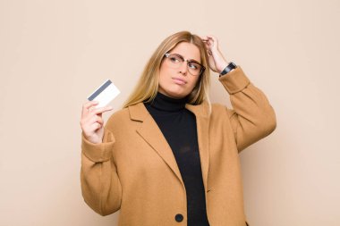 Genç sarışın kadın şaşkın ve kafası karışmış hissediyor. Kafasını kaşıyor ve kredi kartıyla yan tarafa bakıyor.