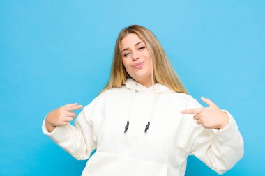 Genç sarışın kadın gururlu, kibirli, mutlu, şaşırmış ve tatmin olmuş görünüyor, kendini gösteriyor, düz duvara karşı kazanan gibi hissediyor.