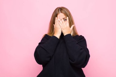 Genç sarışın kadın elleriyle yüzünü kapatıyor, parmaklarının arasından şaşkın bir ifadeyle bakıyor ve düz duvara bakıyor.