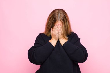 Genç sarışın kadın üzgün, hayal kırıklığına uğramış, gergin ve depresif, iki eliyle yüzünü kapatıyor, düz duvara karşı ağlıyor.