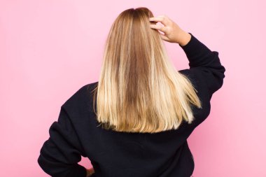 Sarışın genç bir kadın bilgisiz ve kafası karışık, bir çözüm düşünüyor, eli kalçasında, diğeri kafasında, düz duvara karşı dikiz manzaralı.