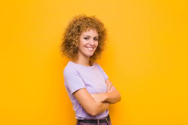 Kameraya çapraz kollu gülümseyen genç afro kadın ve mutlu, kendinden emin, tatmin olmuş bir ifade, turuncu duvara karşı yan bakış.