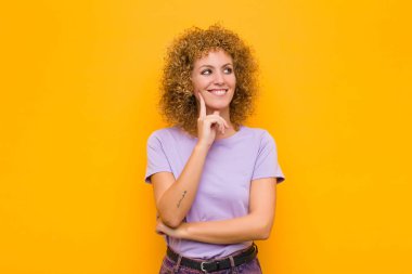 Genç afro kadın mutlu bir şekilde gülümsüyor ve hayal kuruyor ya da şüphe ediyor, turuncu duvara bakıyor.