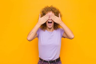 Genç afro kadın gülümsüyor ve mutlu hissediyor, iki eliyle gözlerini kapatıyor ve turuncu duvara karşı inanılmaz bir sürpriz bekliyor.