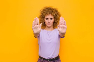 Genç afro kadın ciddi, mutsuz, kızgın ve hoşnutsuz görünüyor. Ya da her iki avuç içi de turuncu duvara yaslıyken dur diyor.