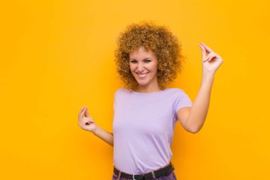 Genç afro kadın gülümsüyor, kaygısız, rahat ve mutlu hissediyor, dans ediyor ve müzik dinliyor, turuncu duvarın karşısındaki bir partide eğleniyor.