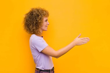 Genç afro kadın gülümsüyor, sizi selamlıyor ve başarılı bir anlaşma için el sıkışmayı teklif ediyor, turuncu duvara karşı işbirliği konsepti