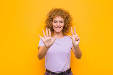 Genç afro kadın gülümsüyor ve arkadaş canlısı görünüyor, 8 ya da 8 numarayı gösteriyor, elini öne uzatmış, turuncu duvara doğru sayıyor.