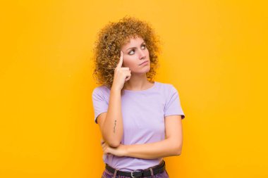 Yoğunlaşmış bakışlı genç bir afro kadın, şüpheli bir ifadeyle, yukarı ve yana bakıyor turuncu duvara karşı