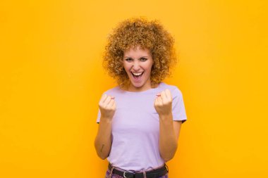 Genç afro kadın zaferle bağırıyor, gülüyor ve turuncu duvara karşı başarıyı kutlarken mutlu ve heyecanlı hissediyor.