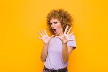 Genç afro kadın korkmuş, geri çekiliyor ve korku ve panik içinde çığlık atıyor, turuncu duvara karşı bir kabusa tepki veriyor.