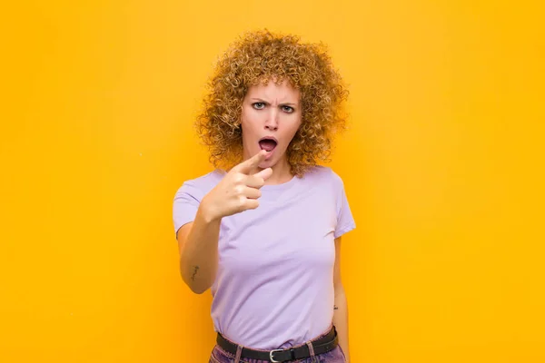 stock image young afro woman pointing at camera with an angry aggressive expression looking like a furious, crazy boss against orange wall