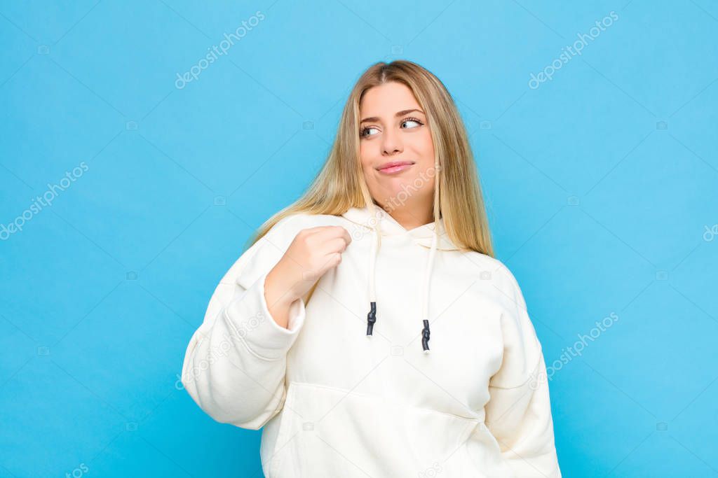 young blonde woman looking arrogant, successful, positive and proud, pointing to self against flat wall