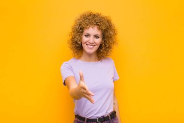 Genç afro kadın gülümsüyor, mutlu, kendinden emin ve arkadaş canlısı görünüyor, anlaşmayı bitirmek için el sıkışmayı teklif ediyor, turuncu duvara karşı işbirliği yapıyor.