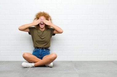 Genç afro kadın gülümsüyor ve mutlu hissediyor, iki eliyle gözlerini kapatıyor ve inanılmaz bir sürprizi bekliyor.