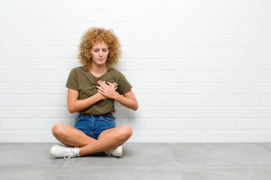 Genç afro kadın üzgün, yaralı ve kalbi kırık görünüyor, iki elini de kalbe yakın tutuyor, ağlıyor ve depresif bir şekilde yerde oturuyor.